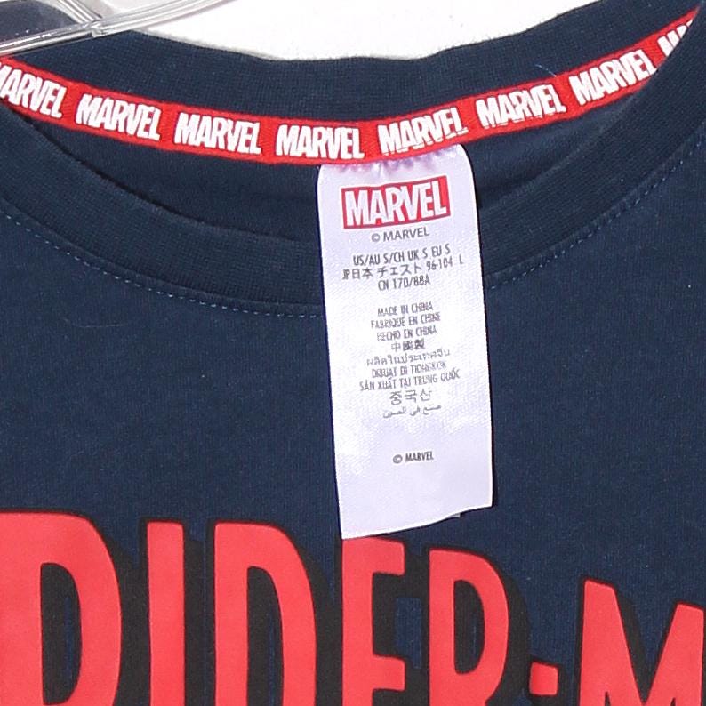 以下が含まれることがあります： 襟に赤いMARVELの文字が入ったネイビーブルーのTシャツ。MARVELのロゴとテキストが入った白いタグが付いています。シャツの下部には大きな赤い「DIDER-M」グラフィックがあります。