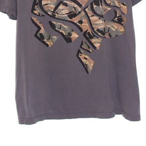 Pode incluir: Uma camiseta cinza com um grande design abstrato na parte superior esquerda. O design apresenta um padr&atilde;o de camuflagem em tons de marrom, verde e cinza, delineado em preto. A camiseta tem mangas curtas e gola redonda.