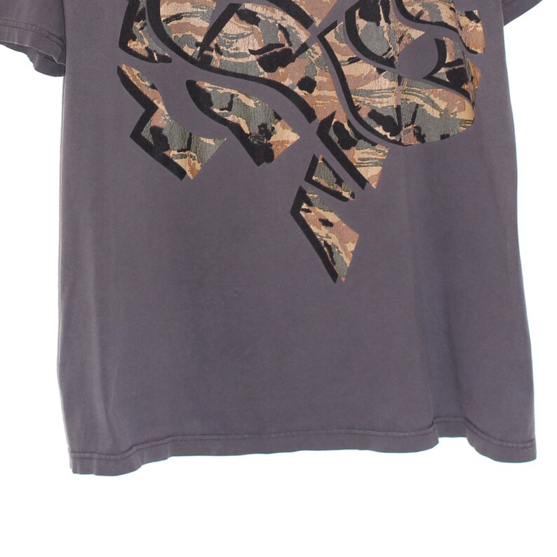 Pode incluir: Uma camiseta cinza com um grande design abstrato na parte superior esquerda. O design apresenta um padr&atilde;o de camuflagem em tons de marrom, verde e cinza, delineado em preto. A camiseta tem mangas curtas e gola redonda.