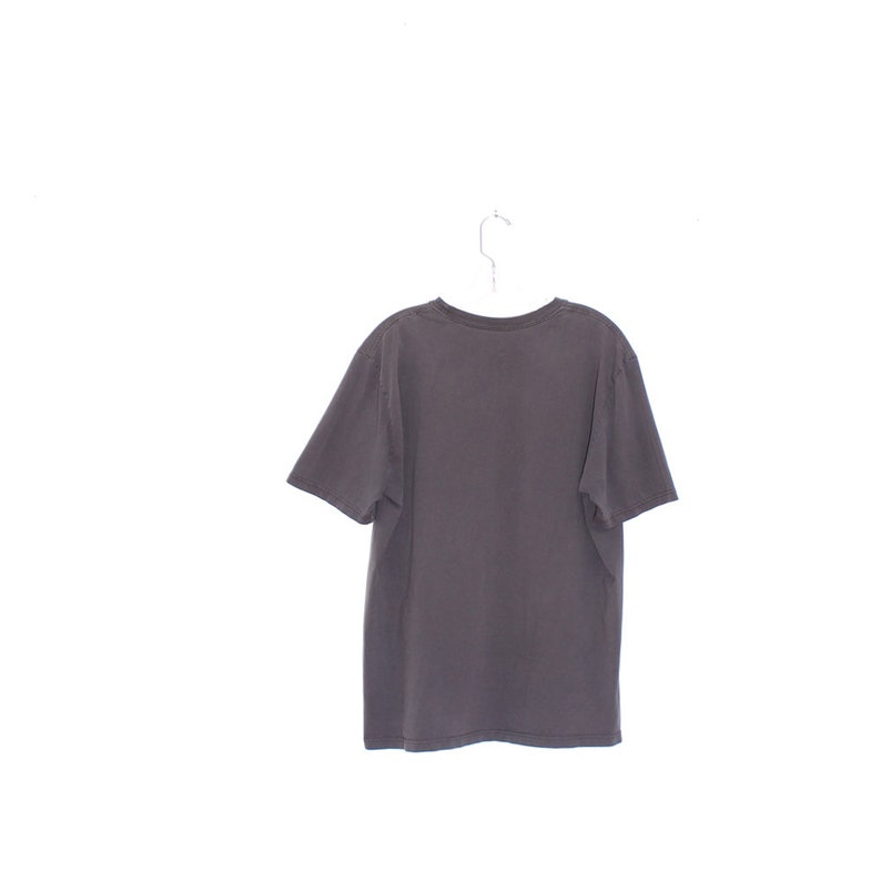 Pode incluir: Uma camiseta cinza escuro pendurada em um cabide de metal contra um fundo branco. A camisa de manga curta tem gola redonda e parece ser feita de um material macio e desgastado. A parte de tr&aacute;s da camisa &eacute; vis&iacute;vel.