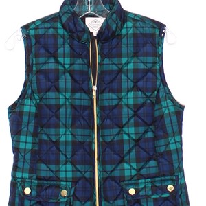 May include: A quilted vest in a green and navy blue plaid pattern. The vest has a gold zipper and two front pockets with gold buttons. The vest is hanging on a clear hanger.