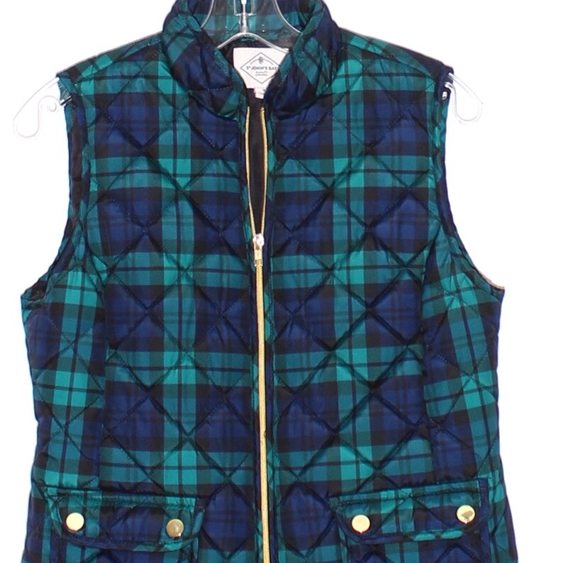 May include: A quilted vest in a green and navy blue plaid pattern. The vest has a gold zipper and two front pockets with gold buttons. The vest is hanging on a clear hanger.