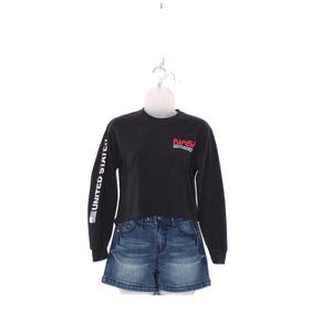 May include: Black long-sleeve shirt with the NASA logo in red and white on the chest. The left sleeve has "UNITED STATES" printed vertically. Paired with denim jean shorts. The outfit is displayed on a white mannequin.