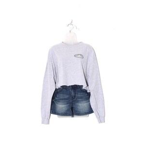 May include: A light gray cropped long-sleeve sweatshirt with a small, oval-shaped design on the chest. Paired with dark wash denim shorts. The outfit is displayed on a white mannequin and hanger.