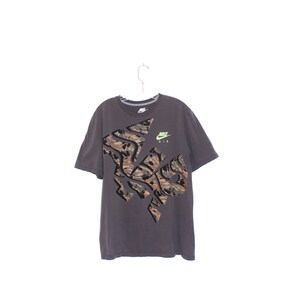 Pode incluir: Uma camiseta Nike cinza escuro com um grande design de camuflagem na frente. A camisa tem mangas curtas e gola redonda. O logotipo da Nike e "AIR" s&atilde;o impressos em verde neon no canto superior direito.