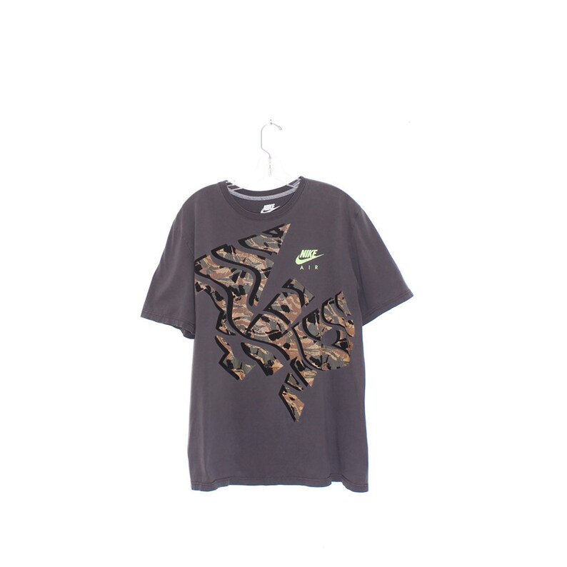 Pode incluir: Uma camiseta Nike cinza escuro com um grande design de camuflagem na frente. A camisa tem mangas curtas e gola redonda. O logotipo da Nike e "AIR" s&atilde;o impressos em verde neon no canto superior direito.