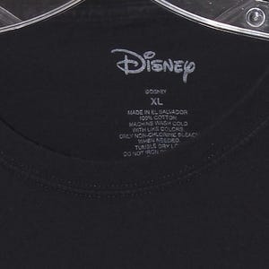 Op de afbeelding: Zwart T-shirt met het witte Disney-logo. Het label geeft maat "XL" en "Made in El Salvador 100% Cotton" aan. Wasaanwijzingen staan op het label. Het kledingstuk hangt aan een hanger.