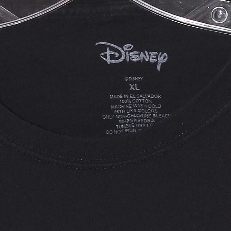 Op de afbeelding: Zwart T-shirt met het witte Disney-logo. Het label geeft maat "XL" en "Made in El Salvador 100% Cotton" aan. Wasaanwijzingen staan op het label. Het kledingstuk hangt aan een hanger.