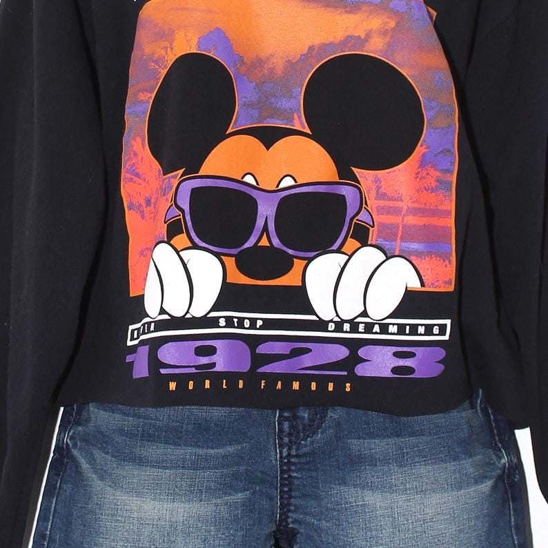 Op de afbeelding: Zwart sweatshirt met een Mickey Mouse-afbeelding met een paarse zonnebril. De tekst luidt "STOP DREAMING 1928 WORLD FAMOUS". Het ontwerp heeft oranje, paarse en witte accenten. Het model draagt een blauwe spijkerbroek.