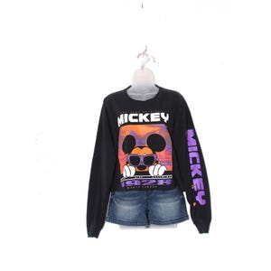 Op de afbeelding: Zwart shirt met lange mouwen met een Mickey Mouse-afbeelding. De afbeelding toont Mickey met een zonnebril tegen een oranje en paarse zonsondergang. De mouw heeft het woord "Mickey" in paars. Gecombineerd met een spijkerbroekshort.