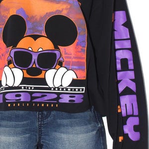 Op de afbeelding: Zwart shirt met lange mouwen met een afbeelding van Mickey Mouse met een zonnebril, tegen een oranje en paarse zonsondergang. De tekst "STOP DREAMING 1928 WORLD FAMOUS" staat eronder. De rechter mouw heeft het woord "MICKEY" in paars.