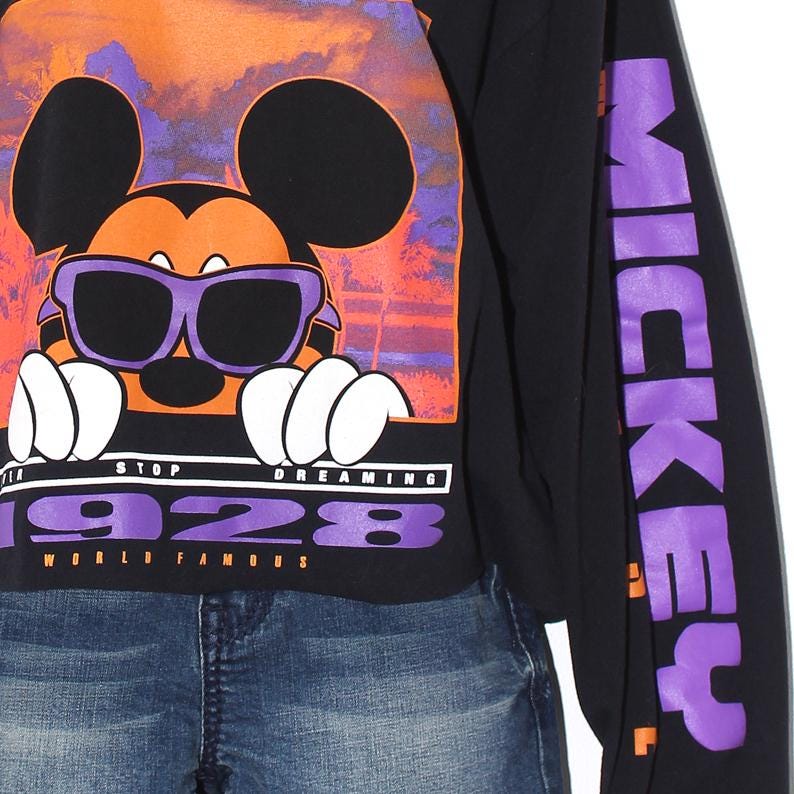 Op de afbeelding: Zwart shirt met lange mouwen met een afbeelding van Mickey Mouse met een zonnebril, tegen een oranje en paarse zonsondergang. De tekst "STOP DREAMING 1928 WORLD FAMOUS" staat eronder. De rechter mouw heeft het woord "MICKEY" in paars.