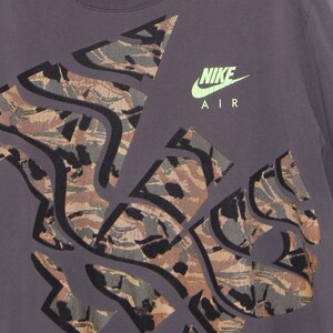Pode incluir: Camiseta cinza escuro com um design de camuflagem em tons de marrom, cinza e preto. O logotipo Nike Air &eacute; verde neon. O design abstrato cobre grande parte da camisa.