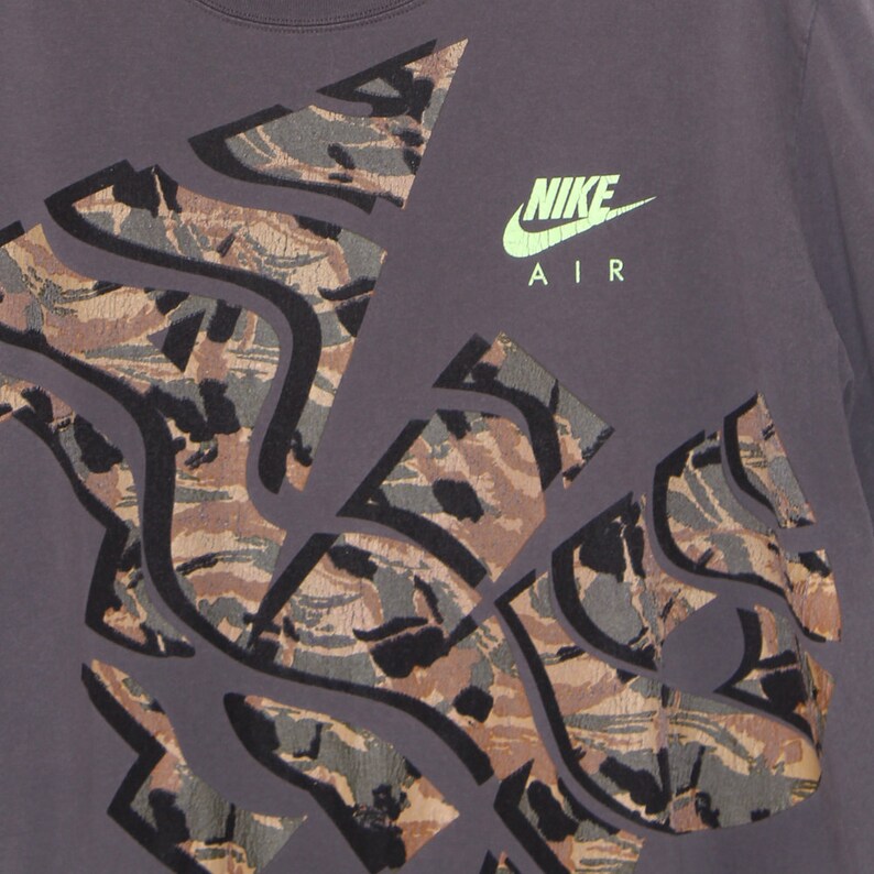 Pode incluir: Camiseta cinza escuro com um design de camuflagem em tons de marrom, cinza e preto. O logotipo Nike Air &eacute; verde neon. O design abstrato cobre grande parte da camisa.