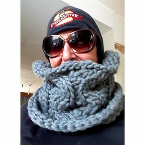 Puede incluir: Una bufanda de punto gris en forma de infinito que se lleva alrededor del cuello de una persona que lleva una boina azul oscuro con un alce y el texto "Kitson & Bitter" bordado en la parte delantera. La persona tambi&eacute;n lleva gafas de sol.