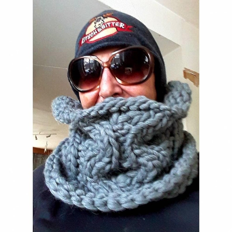 Puede incluir: Una bufanda de punto gris en forma de infinito que se lleva alrededor del cuello de una persona que lleva una boina azul oscuro con un alce y el texto "Kitson & Bitter" bordado en la parte delantera. La persona tambi&eacute;n lleva gafas de sol.