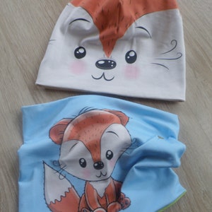 May include: A set of two fox-themed accessories for children. A white beanie with a fox face and a blue neck warmer with a cartoon fox.