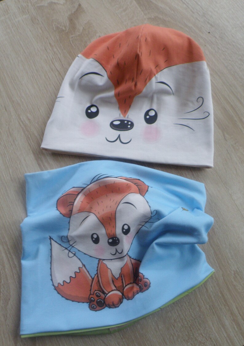 May include: A set of two fox-themed accessories for children. A white beanie with a fox face and a blue neck warmer with a cartoon fox.
