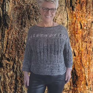 Könnte beinhalten: Eine Frau trägt einen grauen Strickpullover mit einem Used-Look und eine schwarze Lederhose. Sie steht vor einem strukturierten braunen Hintergrund.