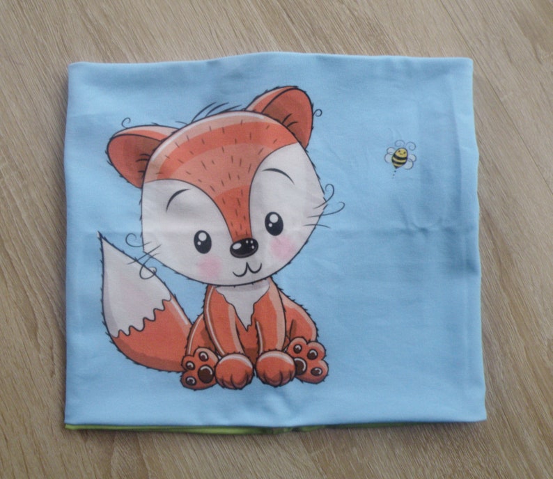 May include: A light blue fabric with a cartoon fox and a bee design. The fox is orange and has a white belly and a fluffy tail. The bee is yellow and black.