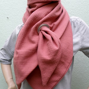 May include: A dusty rose scarf with a textured weave, draped around a mannequin's neck. The scarf is secured with a large, silver-colored ring. The fabric is soft and appears to be lightweight, suitable for cooler weather.