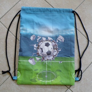 Könnte beinhalten: Eine blaue Sporttasche mit einem Fußball, der durch eine zerbrochene Wand auf einem grünen Feld bricht. Die Tasche hat schwarze Kordeln.
