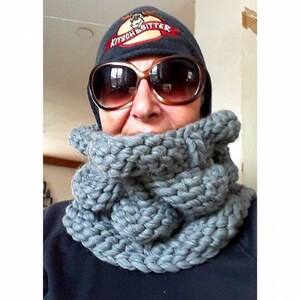 Puede incluir: Una bufanda de punto gris en forma de infinito con una textura gruesa y abultada. La bufanda la lleva puesta una persona que lleva un gorro gris con un logotipo de alce y el texto "Kitsch & Bitter".