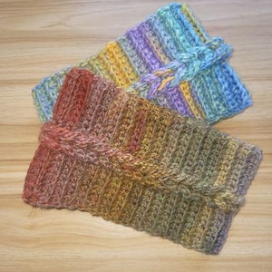 Puede incluir: Dos diademas de ganchillo hechas a mano. Una es una mezcla de azul, morado y amarillo, con un detalle trenzado. La otra es una mezcla de marrón, verde y amarillo. Ambas están dobladas y tienen una textura acanalada.