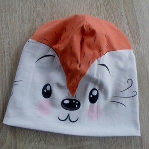 May include: A white and orange knit hat with a cartoon fox face. The fox has black eyes, a black nose, and a pink blush on its cheeks.