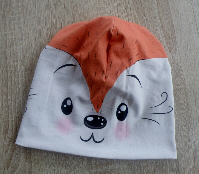 May include: A white and orange knit hat with a cartoon fox face. The fox has black eyes, a black nose, and a pink blush on its cheeks.
