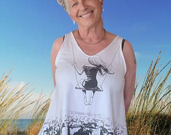 May include: A woman with short, gray hair is wearing a white tank top with a black and white graphic of a girl on a swing. The graphic is set against a cityscape background. The tank top has a scoop neck and a relaxed fit.