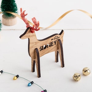 May include: A wooden reindeer ornament with a red and brown antler, the name "Sidney" is cut out of the wood. The ornament is on a white surface with a string of lights and two gold bells.