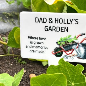 May include: White garden sign with the text "Dad & Holly's Garden" and the phrase "Where love is grown and memories are made". The sign also features a small wheelbarrow with plants and a watering can.