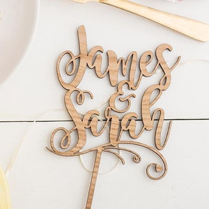 May include: A wooden cake topper with the names "James & Sarah" written in cursive script. The topper is brown and has a simple design.