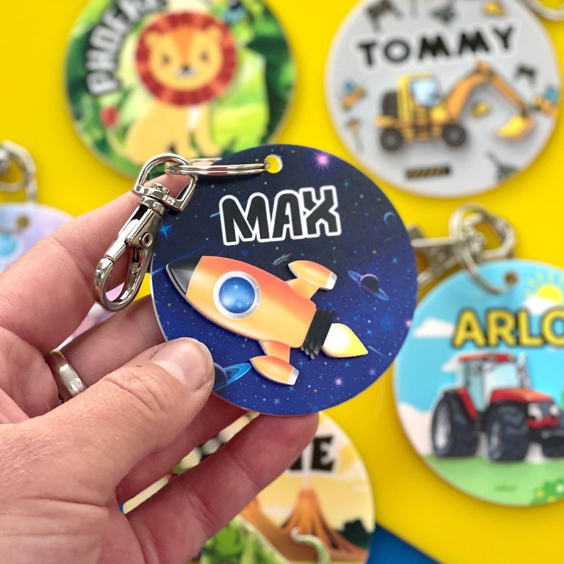 May include: A round, blue and black space-themed keychain with a yellow rocket ship and the name "MAX" printed in white. The keychain has a silver metal ring and clip.