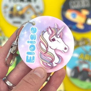 May include: A pink and blue round keychain with a white unicorn with a rainbow mane and horn. The keychain has the name "Eloise" written in blue with white stars.