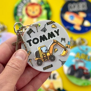 May include: A white round keychain with a yellow excavator, construction tools, and the name "TOMMY" printed in black.