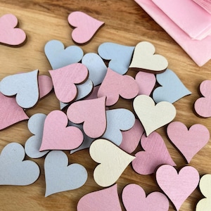 May include: A pile of wooden heart-shaped decorations in pink, blue, and cream colours. The hearts are scattered on a wooden surface.