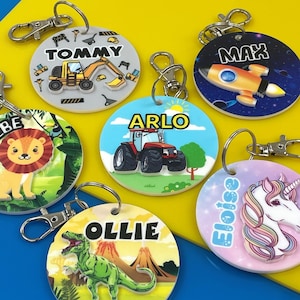 May include: Six colorful round keychains with different designs and names. The keychains feature a tractor, a rocket, a lion, a dinosaur, a unicorn, and a construction vehicle. Each keychain has a silver metal ring.