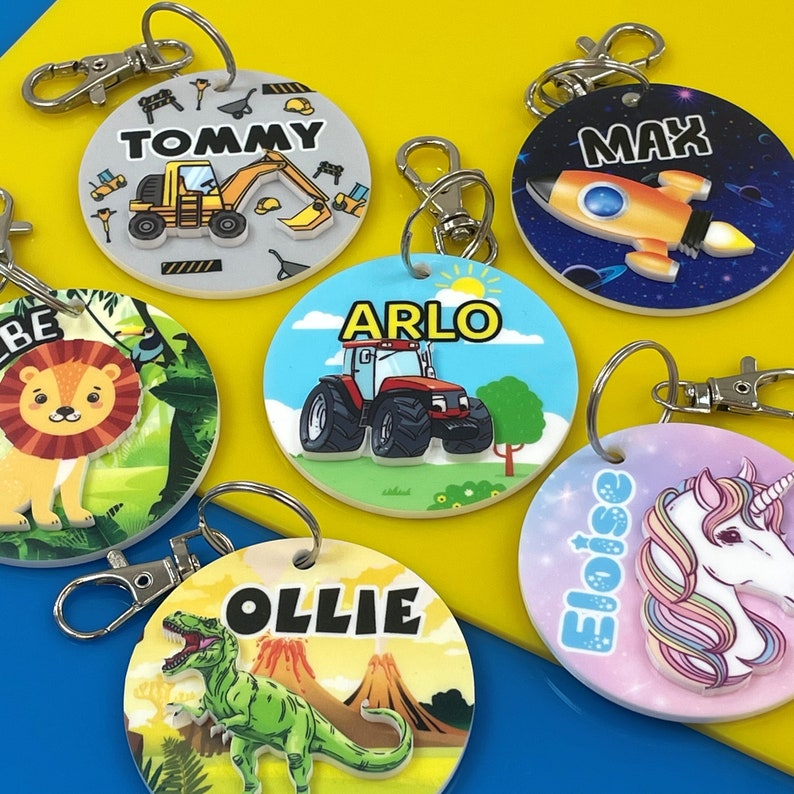 May include: Six colorful round keychains with different designs and names. The keychains feature a tractor, a rocket, a lion, a dinosaur, a unicorn, and a construction vehicle. Each keychain has a silver metal ring.