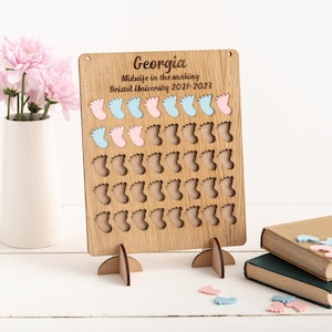 May include: A wooden board with the name "Georgia" and the text "Midwife in the making Bristol University 2021-2023". The board has 49 small footprints cut out, with some filled in with blue and pink footprints. The board is on a stand with two legs.