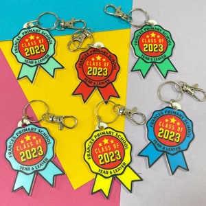 May include: Six colourful keychains with a ribbon design. Each keychain features the text "Frances Primary School" and "Class of 2023" with three gold stars. The ribbons are in different colours: blue, yellow, red, green, and turquoise.