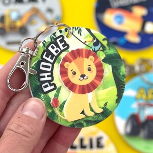 May include: A round, green, and yellow jungle-themed keychain with a lion, toucan, and the name "PHOEBE" printed on it. The keychain has a silver metal clasp.