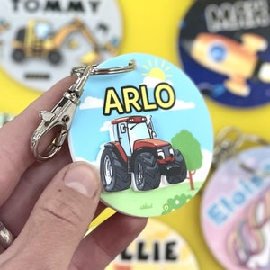 May include: A red and black toy tractor with a yellow sun and blue sky on a round, white, personalized keychain with the name "ARLO" in yellow letters.
