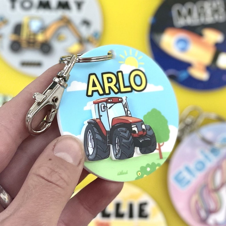 May include: A red and black toy tractor with a yellow sun and blue sky on a round, white, personalized keychain with the name "ARLO" in yellow letters.