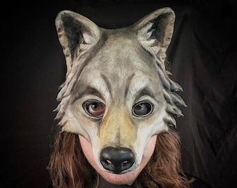 Puede incluir: Una persona con una máscara de lobo gris con ojos negros y nariz negra. La máscara está hecha de fieltro y tiene un aspecto realista. La persona lleva una camisa negra y tiene el pelo largo y castaño.