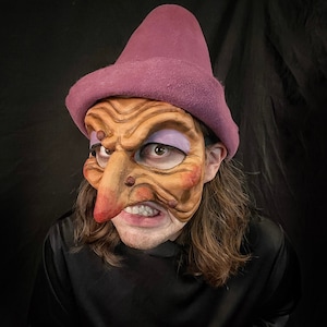 Könnte beinhalten: Eine Person trägt einen lila Hut und eine braune, strukturierte Maske mit einer großen, krummen Nase. Die Maske hat bemalte Details, darunter lila Lidschatten und ein breites, zahniges Grinsen.