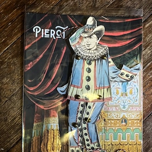 May include: A vintage Pierrot paper doll with a blue and cream costume, a ruffled collar, and a hat. The doll is set against a backdrop of a stage curtain and ornate architectural details. The word "Pierrot" is written at the top.
