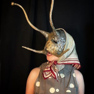 Könnte beinhalten: Eine einzigartige, handgefertigte Maske mit metallischer, strukturierter Oberfläche und länglichen Antennen mit knollenförmigen Spitzen. Die Maske wird mit einem Kopftuch und einem gepunkteten Kleid getragen, was ein surreales und künstlerisches Kostüm ergibt.