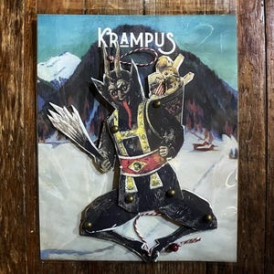 May include: A Krampus-themed paper cutout decoration. The black figure has horns, a long tongue, and carries a basket with children. The background features a snowy mountain scene. The word "KRAMPUS" is written in white.